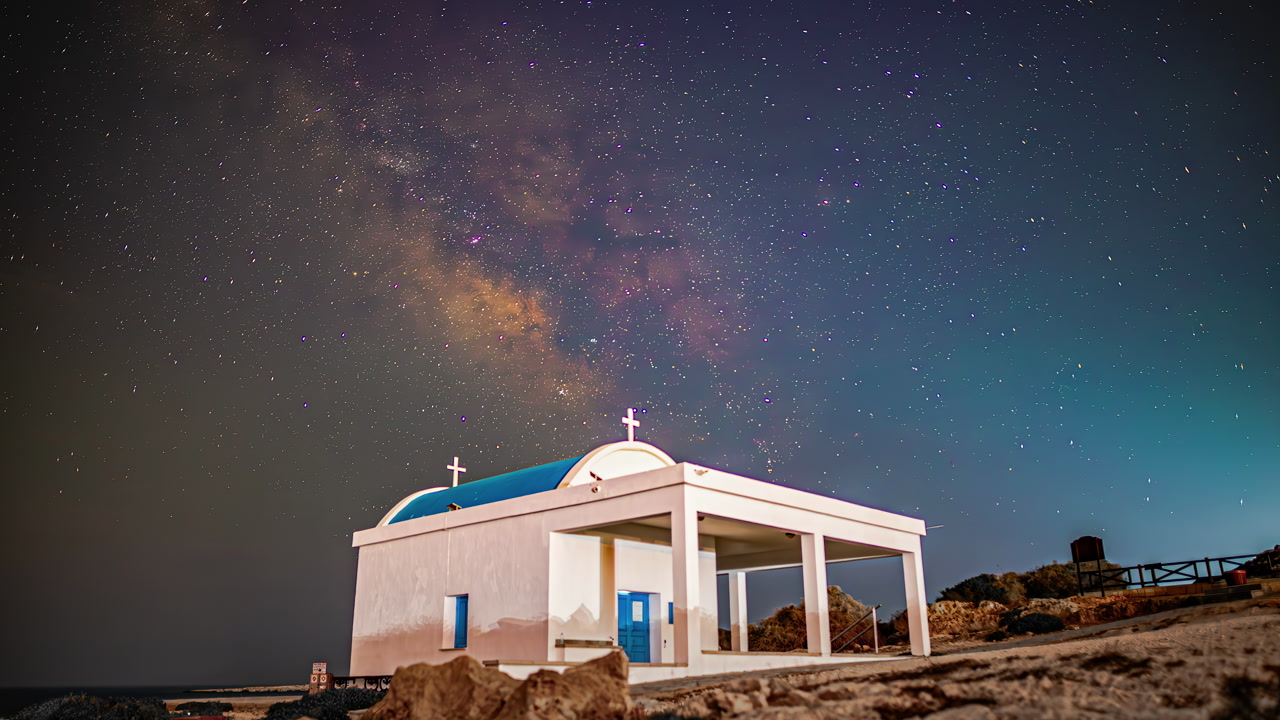Stunning night sky time-lapse captures Milky Way galaxy core rotating above iconic white-washed Ayioi Anargiroi Orthodox chapel with blue dome on rocky Cape Greco Cyprus coastline