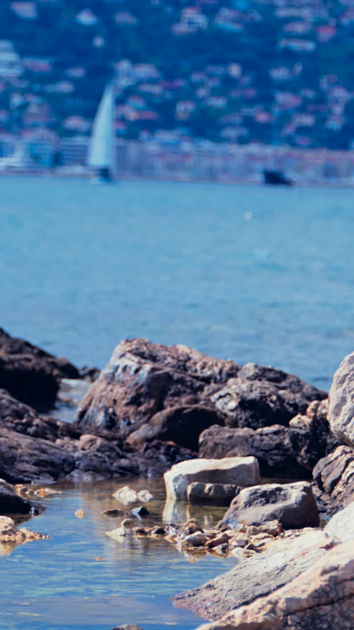 Rocce sulla riva con una barca a vela che si muove sul mare a Juan-les-Pins, in Francia, con la città e la montagna sullo sfondo