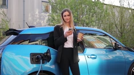 a woman stands by an electric car, watching her phone as it charges. she is focused on the charging process, multitasking with her mobile device
