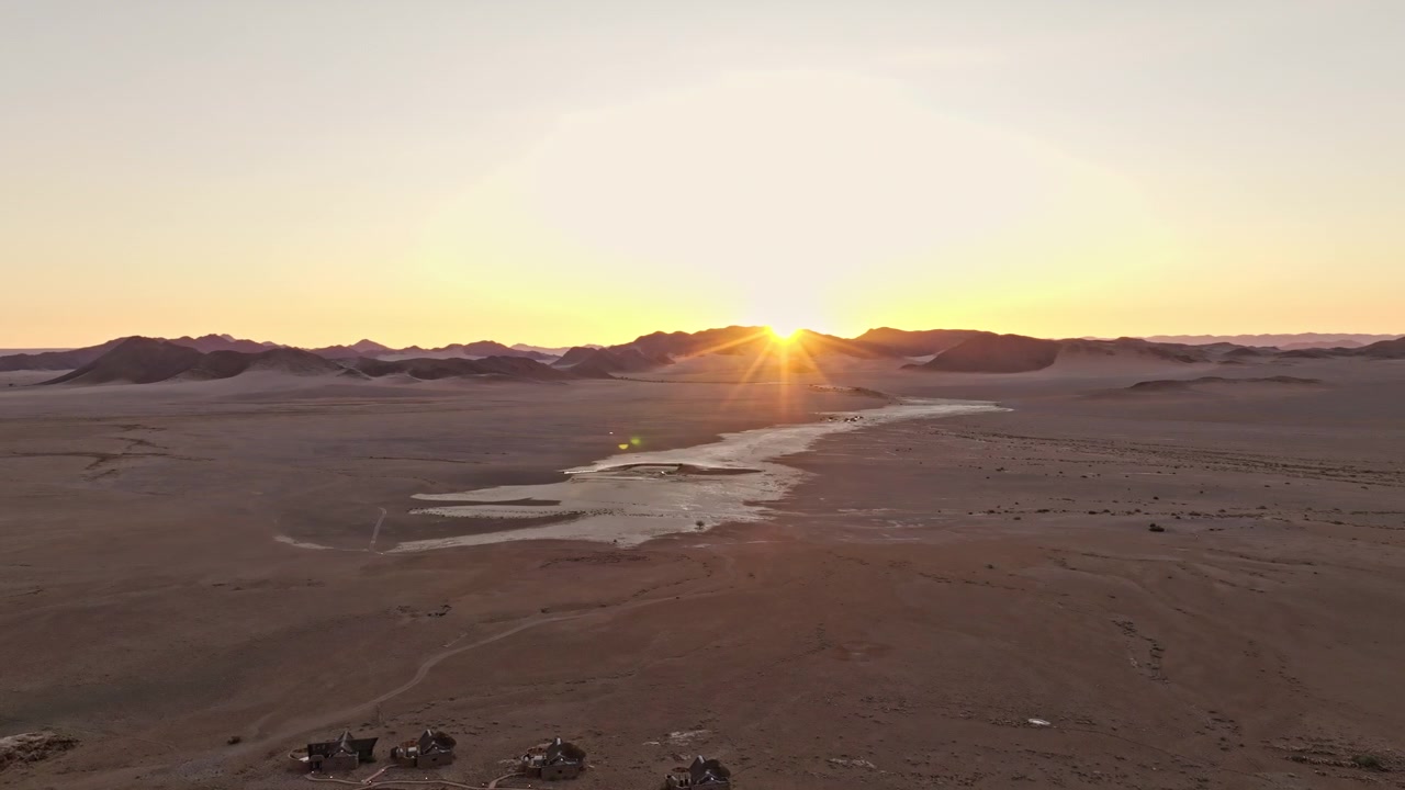 Drone-näkymä pehmeästä auringonlaskun hehkusta laajan aavikkotasangon ja kuivan uoman yllä Namibiassa. Lämmin matala valo pyyhkii yli villien soratasankojen ja kaukana olevien vuorten rauhallisessa elokuvamaisessa tunnelmassa.