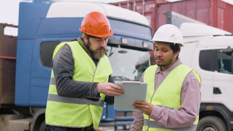 Boss-And-Worker-Wearing-Vests-And-Safety-Helmets-Organizing-A-Truck-Fleet-In-A-Logistics-Park-While-They-Consulting-A-Document-1