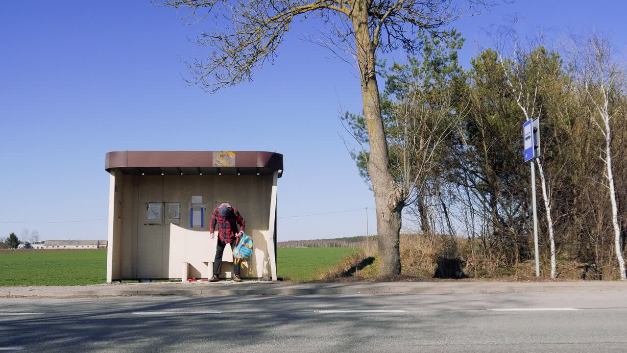 A young man waits for a bus at an old, rural bus stop. The bright, clear sky and surrounding nature emphasize the solitude of the moment.