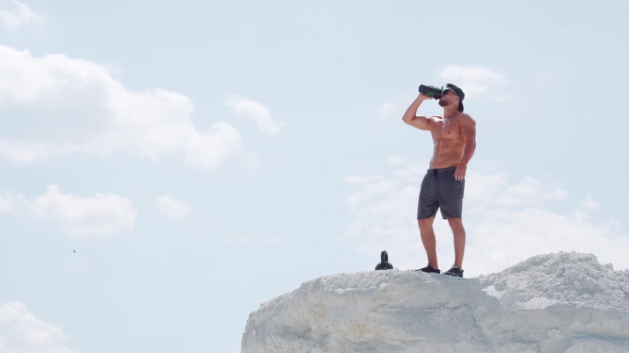 Strong man drinks water from a bottle in an outdoor training session. Athlete on a white mountain against the blue sky