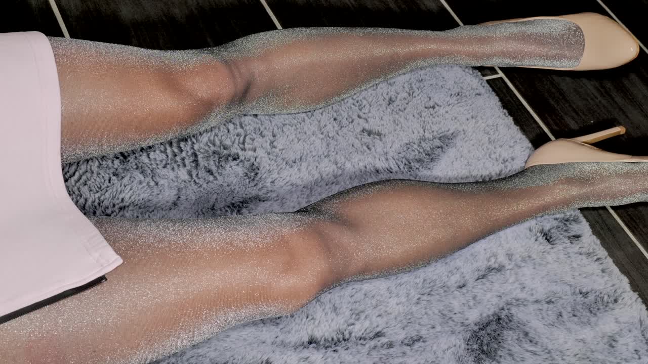 미니스커트와 아름다운 긴 다리를 가진 반짝이는 스타킹을 입은 여성은 검은 가죽으로 만든 하이힐 부츠를 신고 바닥에 앉아 초조하게 다리를 앞뒤로 움직입니다.