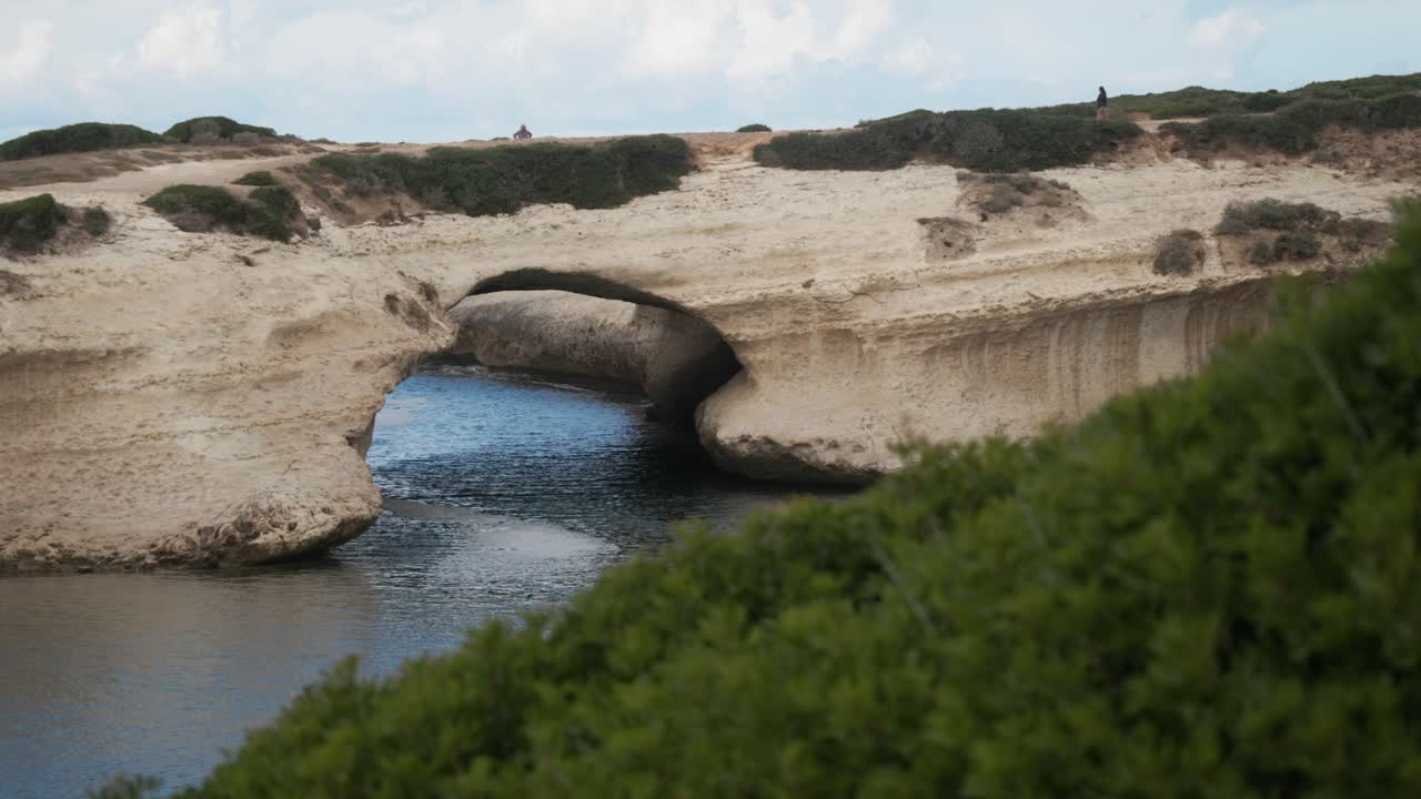 S'Archittu's natural rock arch in Sardinia, Italy. Waves crash against the beach, showcasing a stunning coastal landscape. Perfect for travel, nature, and scenic beach footage.