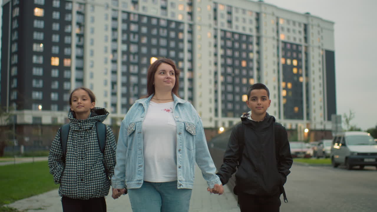 Confident woman walks with two schoolchildren holding hands in city area near tall residential buildings during evening, showing strength, unity, and support in urban family life with backpacks and winter clothes
