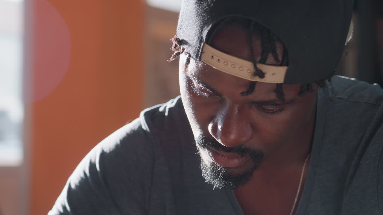 Close up of young man wearing cap writing notes in book while studying at library desk, surrounded by stacks of textbooks, focused expression under warm sunlight