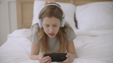 joyful-teen-girl-is-reading-text-on-screen-of-mobile-phone-lying-on-bed-using-wireless-headphones-and-wifi