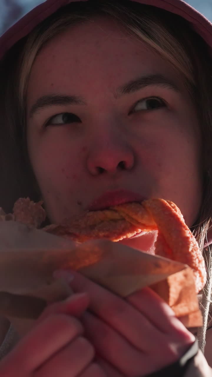 primer plano de una mujer joven de pie al aire libre, comiendo un bocadillo mientras deja caer comida para los pájaros en la casa de pájaros de un parque nevado, luces de la calle suaves brillando en el fondo
