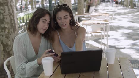 young female friends sitting in city cafe and using laptop