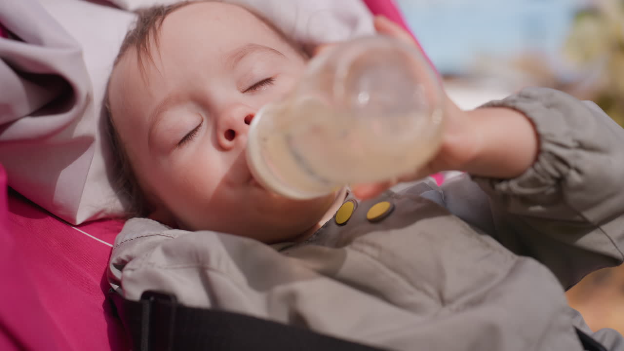 Bebê bebendo mamadeira no carrinho, cena aproximada em parque ao ar livre, bebê sonolento descansando em carrinho rosa, luz solar suave, rotina de alimentação tranquila, passeio familiar calmo, jaqueta e cobertor aconchegantes