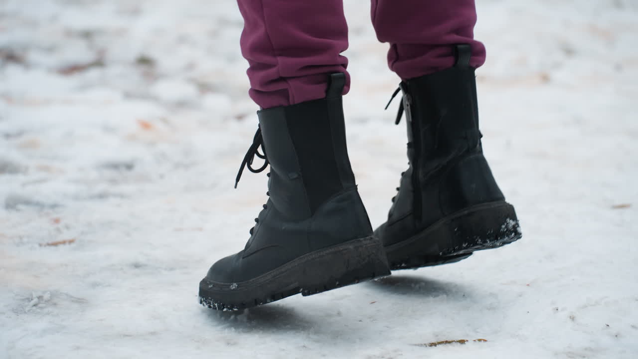 vista lateral de un individuo con botas negras saltando juguetonamente en el suelo cubierto de nieve con follaje seco esparcido, el entorno de invierno captura el movimiento, el calzado y la textura del terreno helado