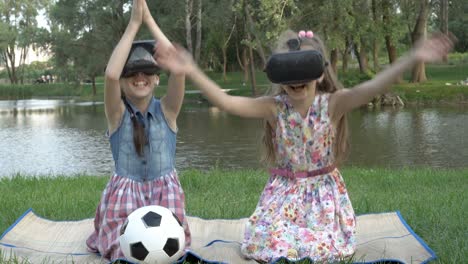 two fun little girls play a virtual game together while sitting in virtual reality glasses on the grass in a city park at sunset. close-up. the concept of virtual games. 4k.