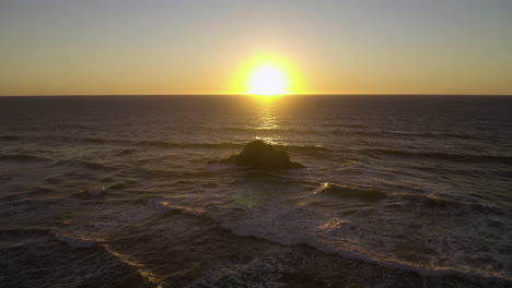 sun sets on horizon directly above single rock in ocean battered by waves as spray rises, oregon coast