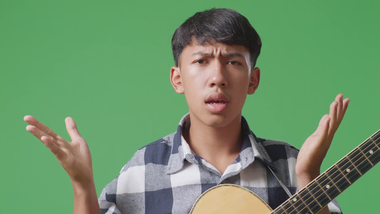 기타를 들고 있는 아시아 청소년의 클로즈업은 당황스러운 몸짓을 보여주고 녹색 스크린 배경에서 무엇을 말하고 있습니다.