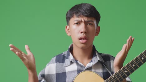 primer plano de un adolescente asiático con una guitarra mostrando un gesto desconcertado y diciendo qué en el fondo de la pantalla verde
