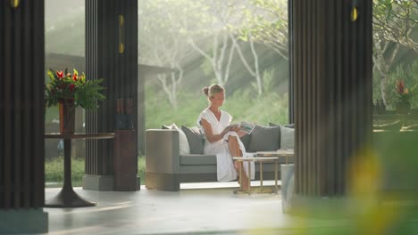 woman in white dress sitting in luxury open resort lounge reading with morning light rays