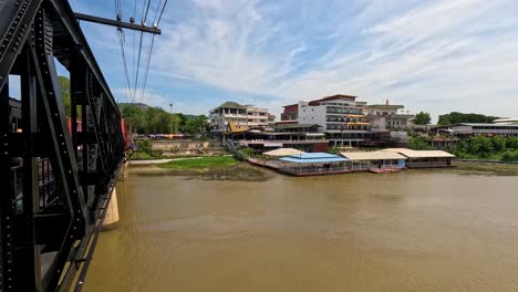 camera moves smoothly across a steel bridge, revealing riverside buildings and the wide river kwai under bright daylight with partly cloudy skies