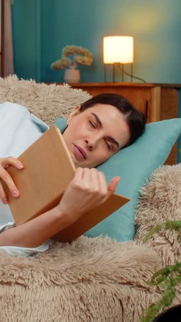 young woman falling asleep lying on sofa peacefully holding open book tired after reading relaxing