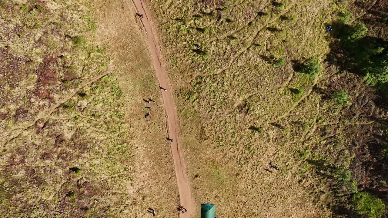 Top-down aerial view of a dirt hiking trail running through a grassy mountain field with colorful tents and scattered hikers enjoying the outdoors