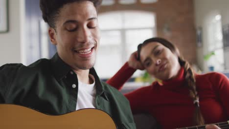 happy biracial couple sitting on sofa in living room playing guitar