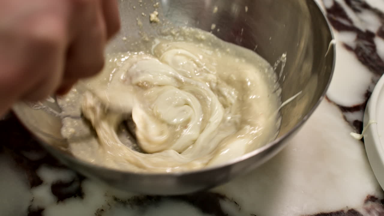 close-up shot of a chef's hands whisking a smooth, creamy white sauce in a stainless steel bowl. Professional culinary preparation in a generic kitchen setting