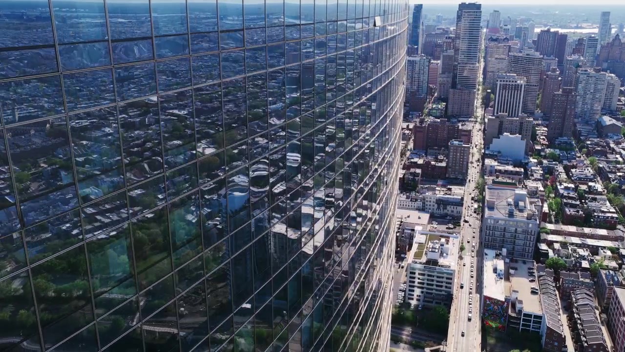Vista aérea de drone da fachada de vidro reflexiva de um arranha-céu da Filadélfia, espelhando a paisagem urbana, o Rio Delaware e os bairros circundantes abaixo.