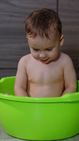 beautiful caucasian toddler sitting in the washbowl smiling happily. lovely kid enjoys his game with the splashing water. vertical video