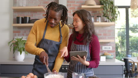 Happy-diverse-couple-in-aprons-using-tablet-and-baking-in-sunny-kitchen,-slow-motion