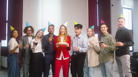 portrait of a group of multiethnic colleagues looking at camera, posing with a birthday cake and raising a toast at the office party