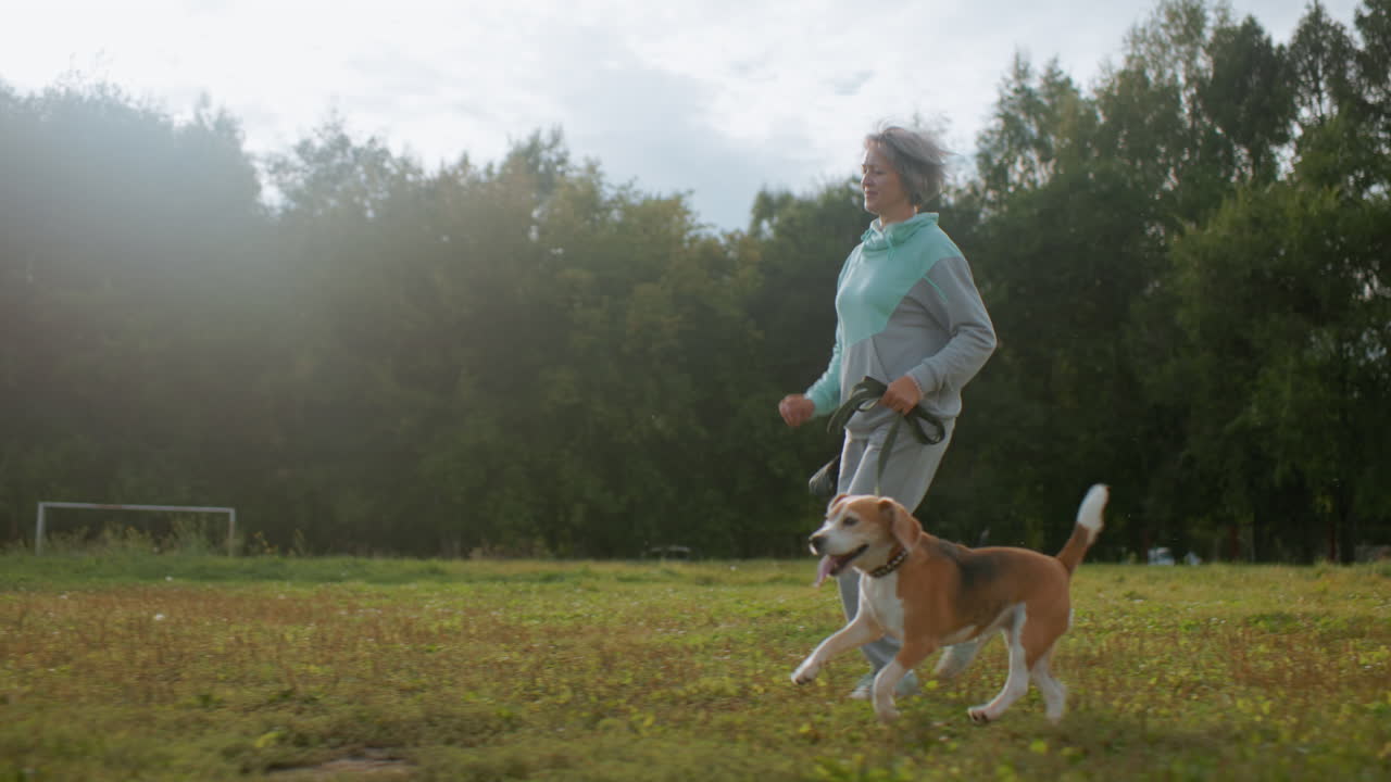 Female athlete jogging with playful puppy across grassy field near table tennis court, cars moving in background under cloudy sky, outdoor training filled with energy, companionship, and fresh air