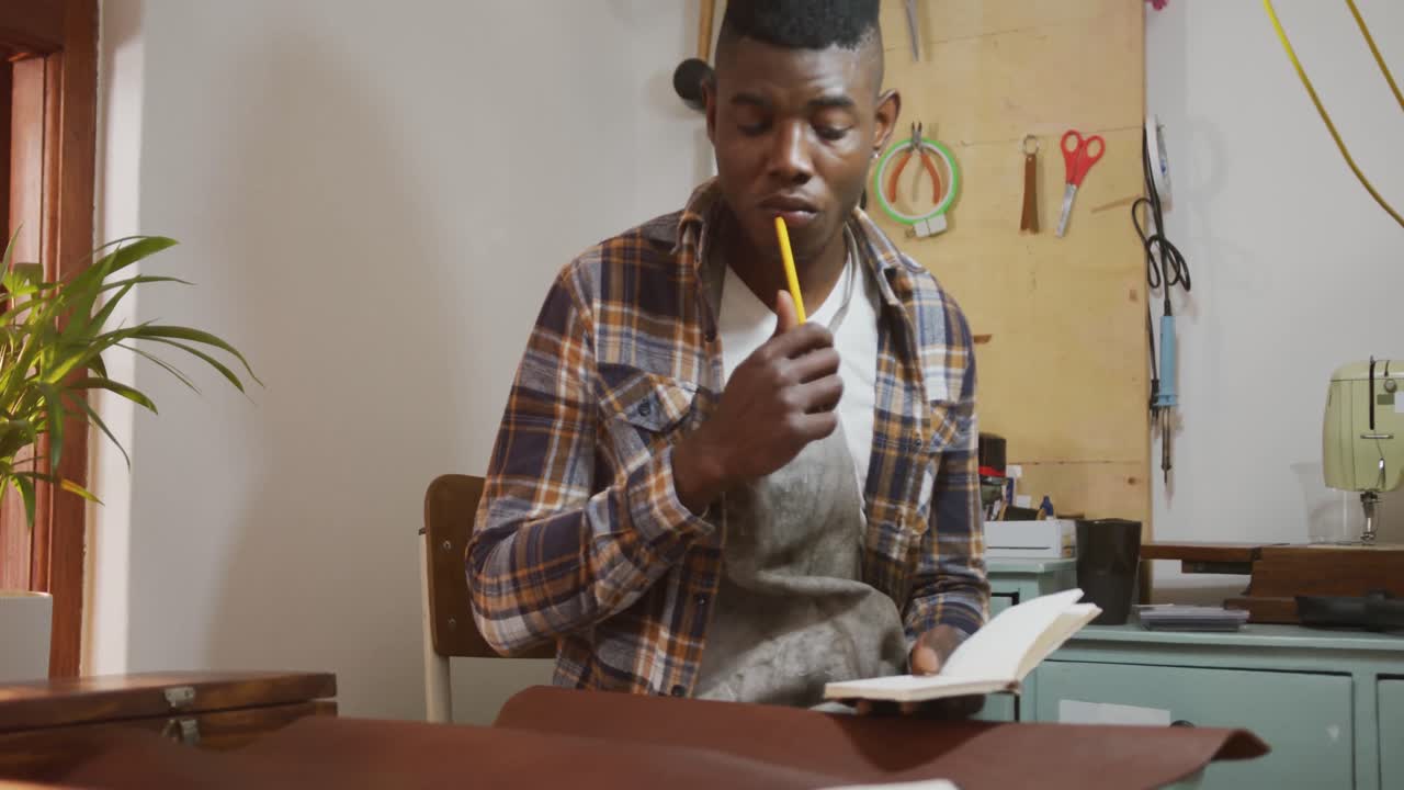 Artesão afro-americano pensativo usando avental fazendo anotações em uma oficina de couro.