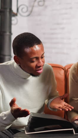 attractive emotional woman tells family stories to black man friend looking pictures in photo-album sitting on couch in living room slow motion