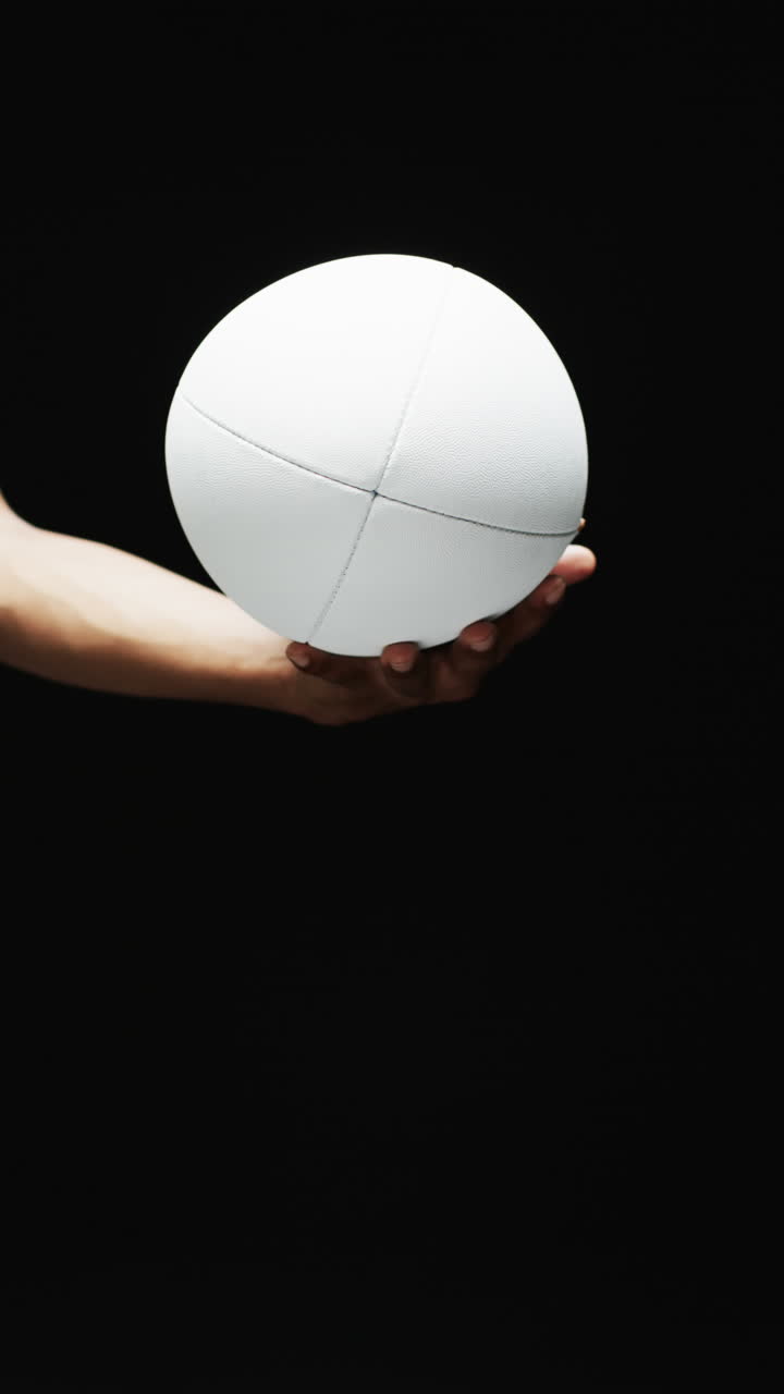Vertical video of hand of african american man holding white rugby ball on black background