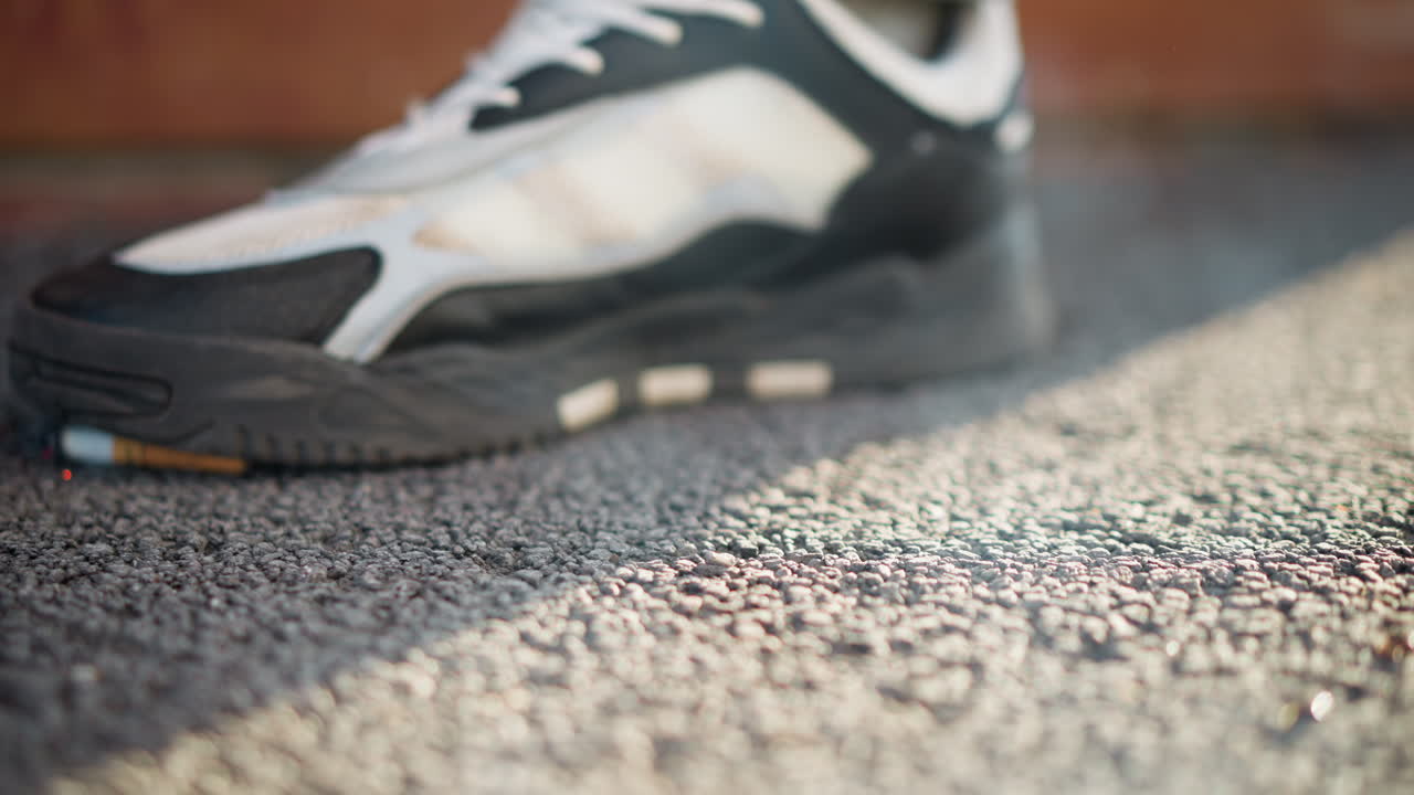 burned cigarette falls to asphalt as man lifts foot to step on it with smoke rising, shallow focus on pavement texture and motion indicating act of extinguishing cigarette in outdoor