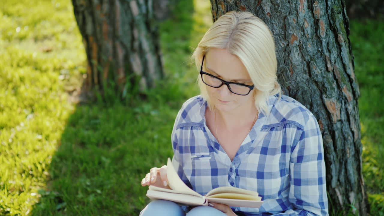 A Blonde Young Woman In Glasses Reads A Book In The Park Sits Near A Tree Beautiful Light Before Sun