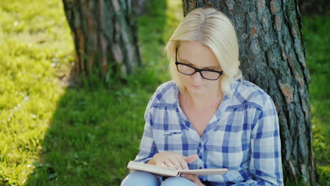 Eine-Blonde-Junge-Frau-Mit-Brille-Liest-Ein-Buch-Im-Park-Sitzt-In-Der-Nähe-Eines-Baumes-Schönes-Licht-Vor-Der-Sonne-Beautiful