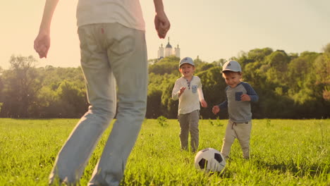 Un-Joven-Padre-Con-Una-Camiseta-Blanca-Con-Dos-Hijos-Jugando-Al-Fútbol-En-El-Césped-Al-Atardecer-Bajo-El-Sol-En-Cámara-Lenta