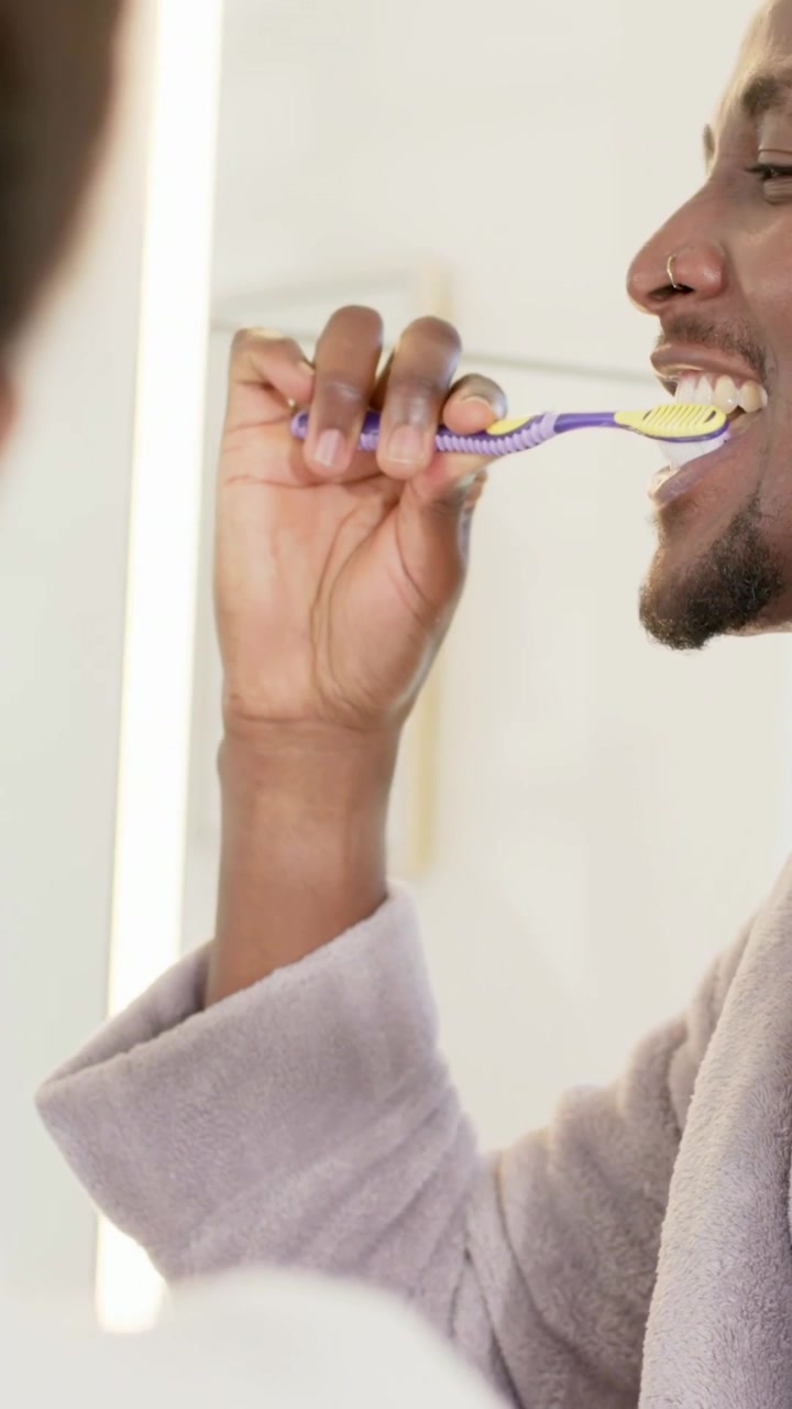Vertical video: Putting brush, man brushing teeth in bathroom wearing gray robe with foam for care