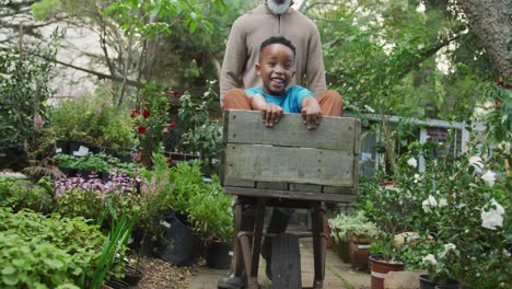 Glücklicher-älterer-Afroamerikanischer-Mann-Mit-Seinem-Enkel,-Der-Mit-Schubkarre-Im-Garten-Spielt
