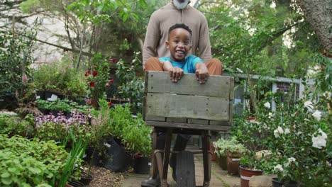 happy senior african american man with his grandson playing with wheelbarrow in garden