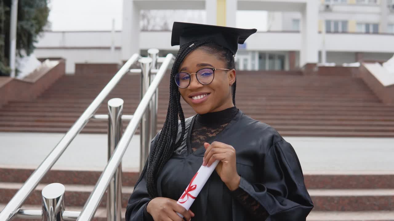 una mujer joven en la universidad en el manto del maestro sostiene un diploma y corrige sus gafas mientras mira a la cámara. retrato frente a la entrada central de la universidad en segundo plano