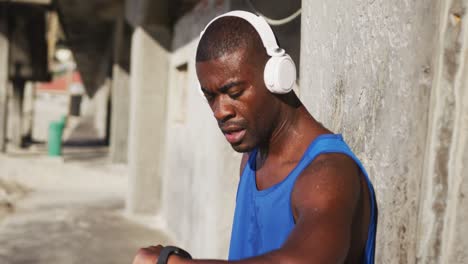 african american man wearing headphones checking his smartphone, exercising outdoors on sunny day