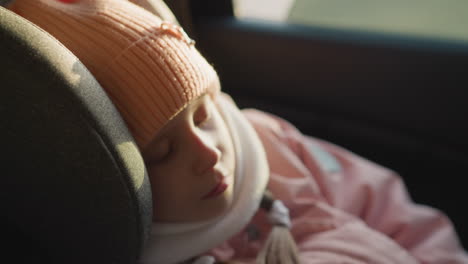 a young girl peacefully sleeps in a car seat, her face softly lit by warm sunlight. she wears a pink knit hat and scarf, embodying tranquility and comfort during a sunny winter day