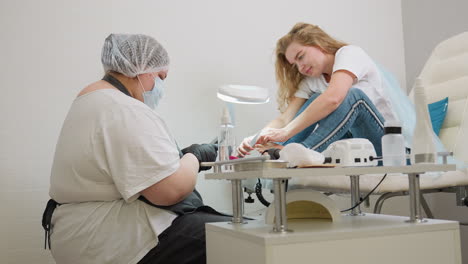 client sitting on spa chair filing big toenail while smiling and engaging with technician in mask and gloves, with pedicure tools and equipment placed on table in bright clean salon setting