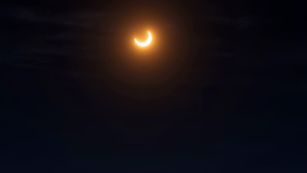 Crescente brilhante permanece perto do centro enquanto finas faixas de nuvens se movem por um céu quase preto, enquanto uma névoa âmbar suave envolve o sol eclipsado e enfatiza sua borda curva iluminada.