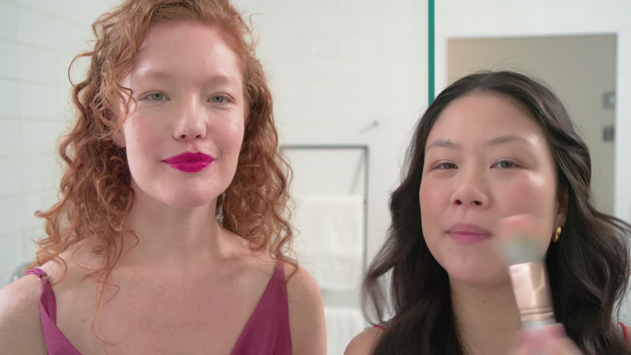 Diverse female friends prompted by camera applying lip with wand and showing blush brush in mirror