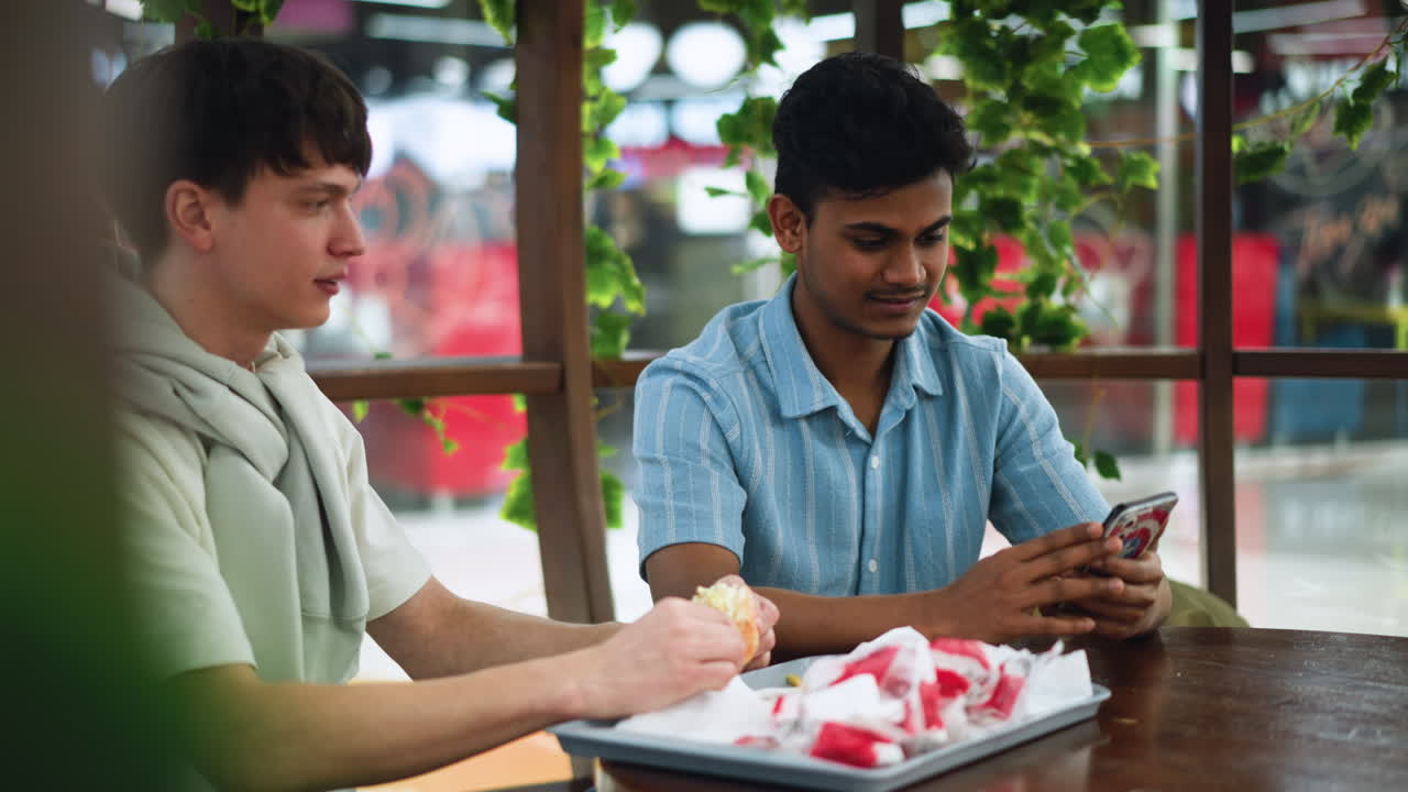 Indian looking man pressing phone screen while peer eats burger beside him, candid cafeteria moment showing interaction, focus on hand movement and food consumption, casual conversation vibe