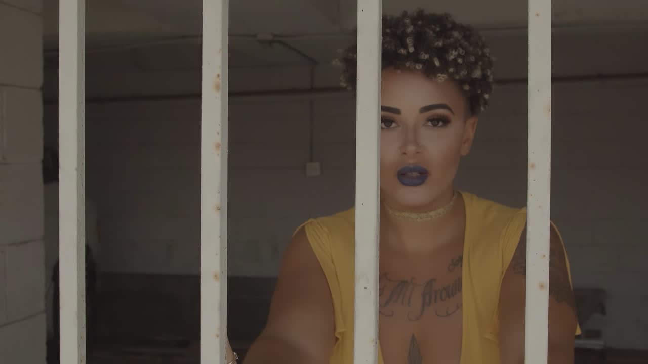 mujer de raza mixta asustada con lápiz labial oscuro y tatuajes tras las rejas en la celda de la prisión, de cerca, de mano