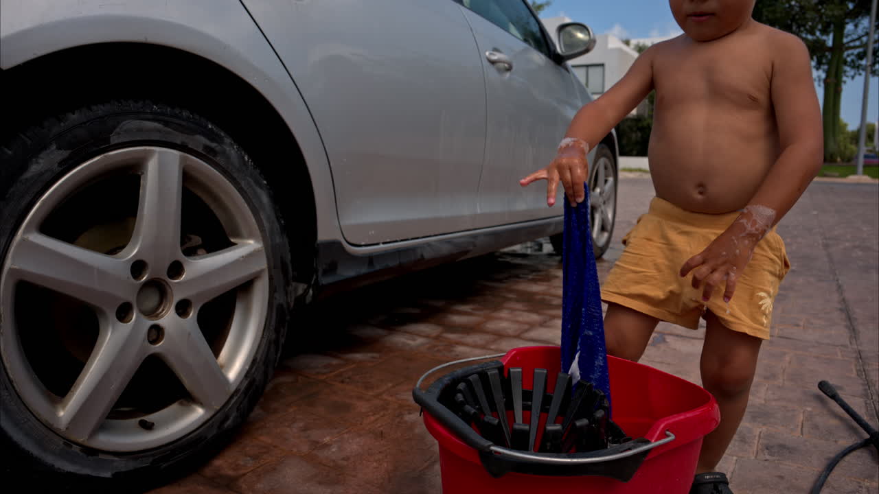 divertido niño latino mexicano sin camisa con pantalones cortos amarillos lavando el coche de plata de la familia con un paño azul empapado en agua jabonosa